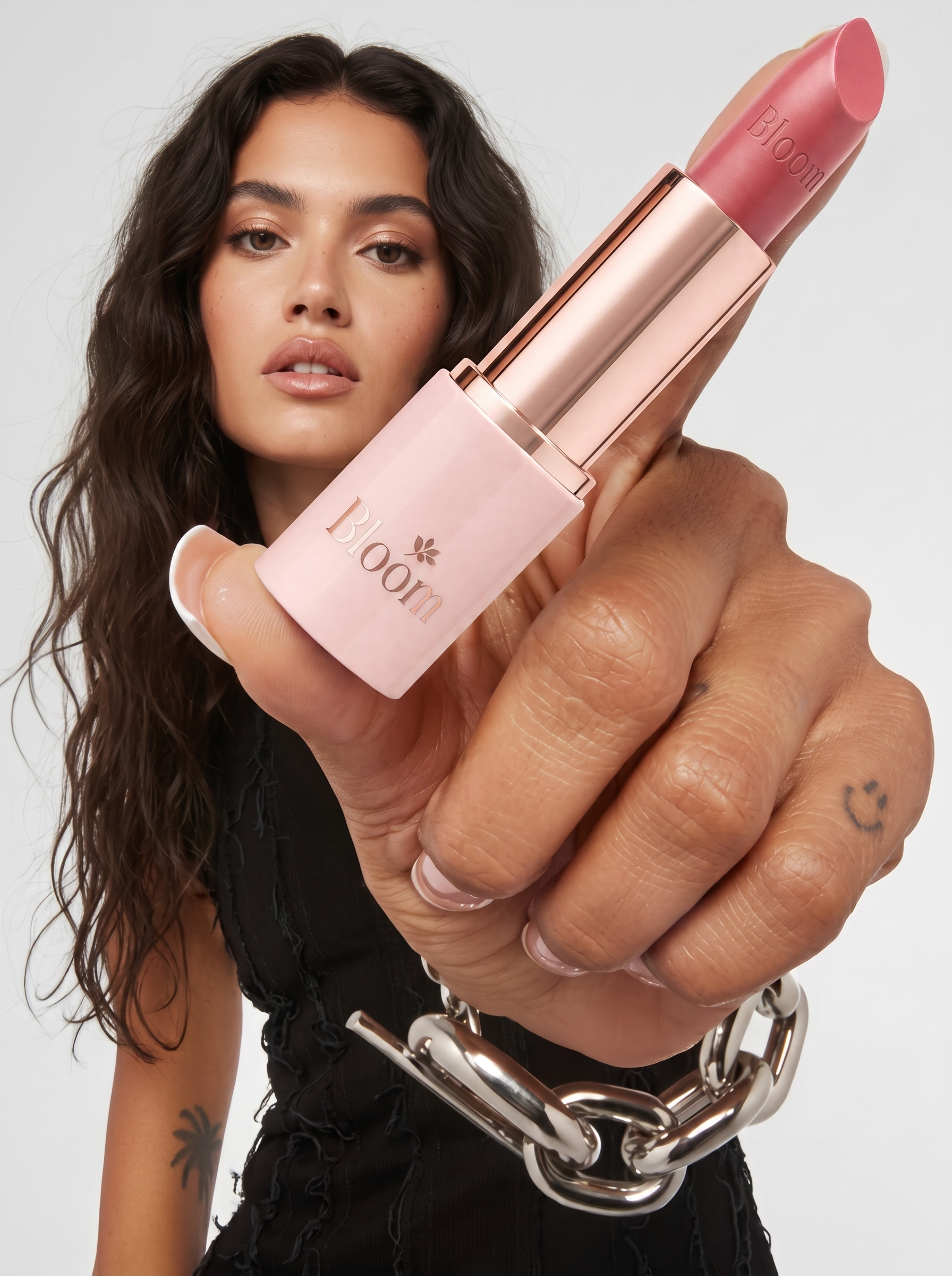 Editorial close-up: a hand holding a rose-gold lipstick towards camera, model out of focus behind